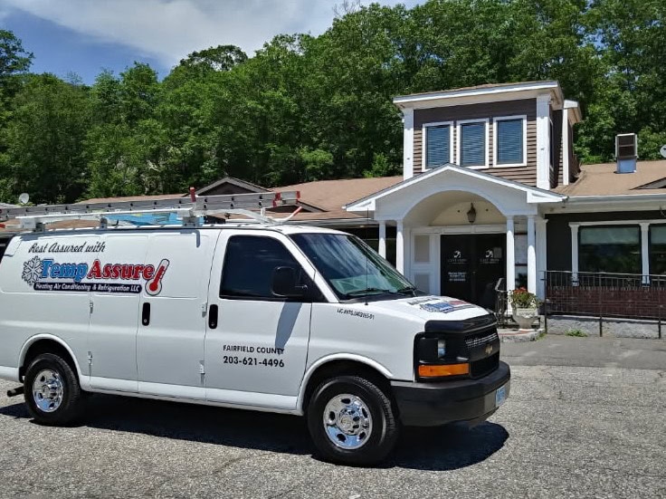 A white Temp Assure service van featuring HVAC Services Fairfield County, CT is parked in front of a building with brown and white exterior walls, surrounded by trees on a sunny day. The van displays company contact information and a roof ladder.